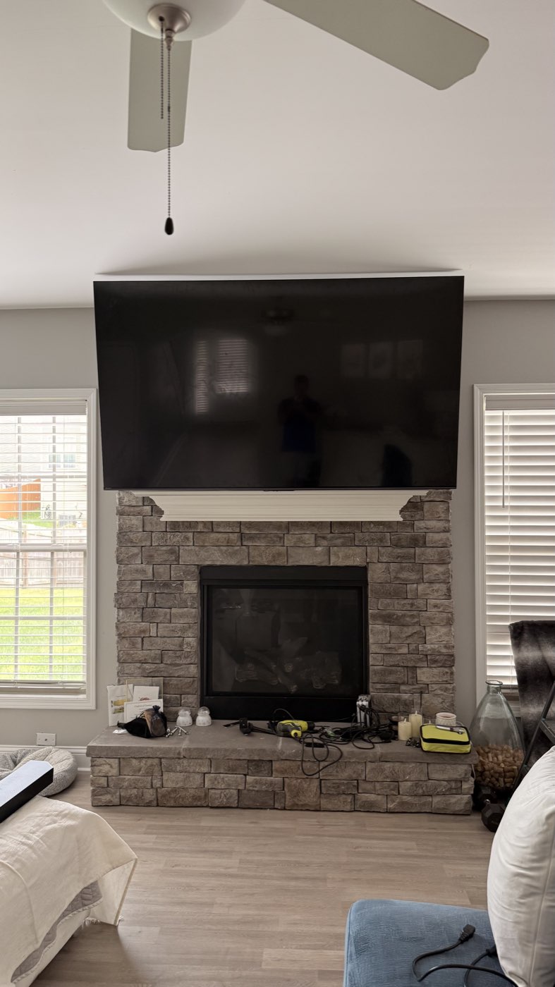 TV mounted above a gray stacked stone fireplace with a white mantel shelf in a living room, mid installation with tools and cables still on the hearth