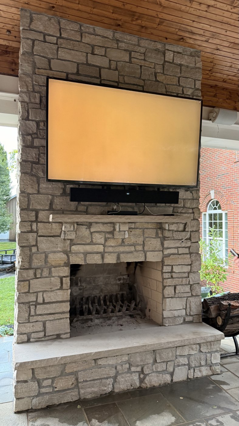 TV and soundbar mounted above a stacked stone fireplace on a covered outdoor patio, with a wood plank ceiling above and a brick house visible in the background