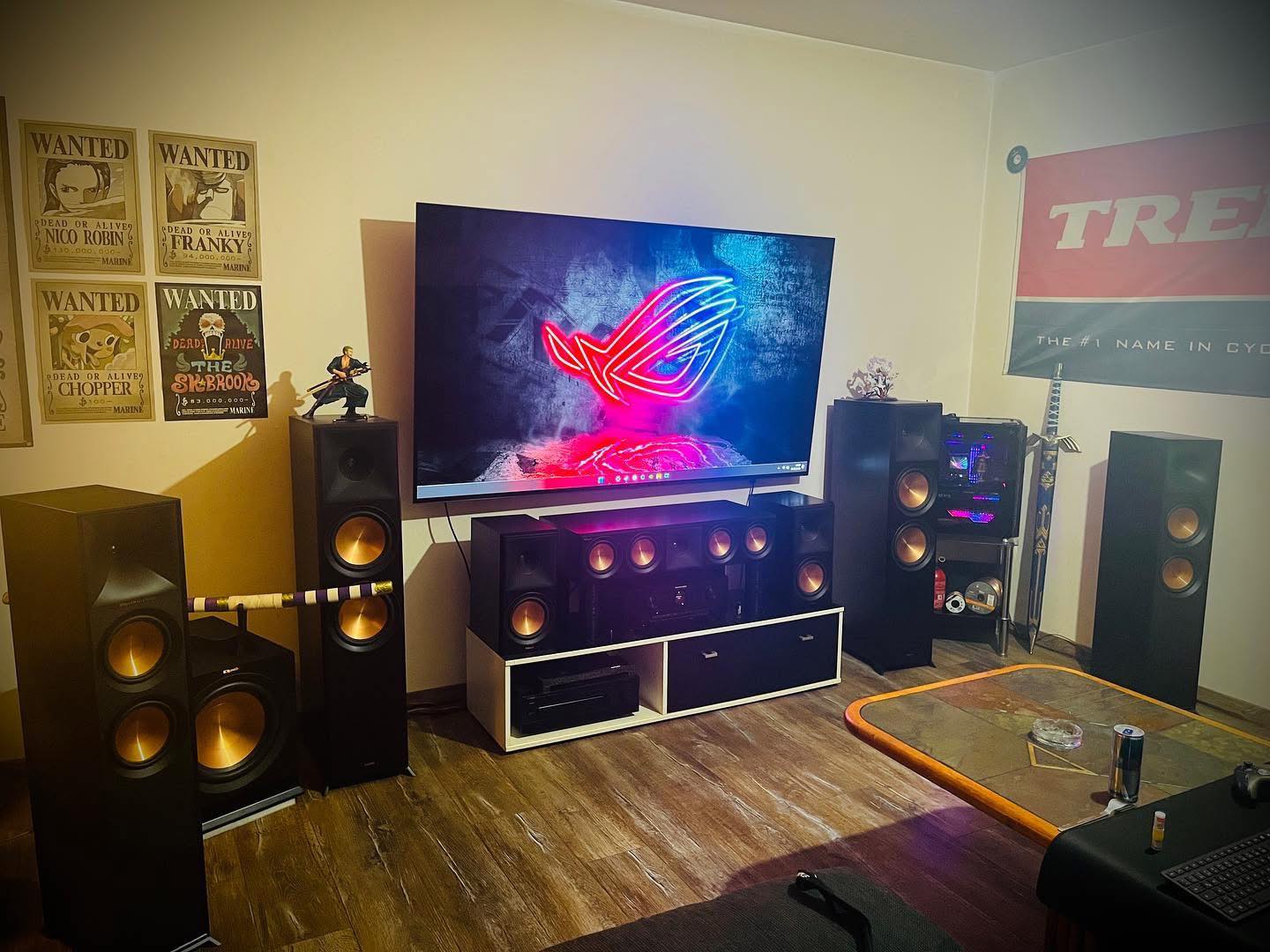 Wall mounted TV in a gaming room above a media unit, with Klipsch tower speakers either side, a center channel, subwoofer and a lit PC tower