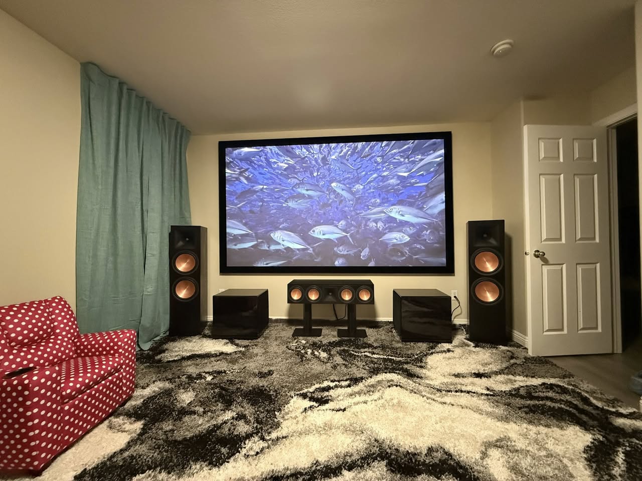 Fixed frame projector screen mounted on a living room wall showing an underwater scene, with Klipsch tower speakers, a center channel and two subwoofers below it
