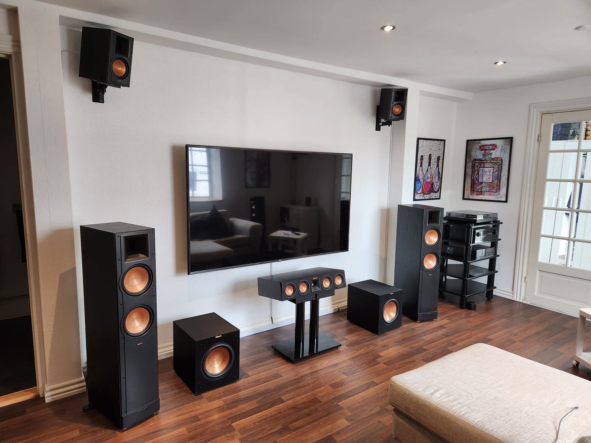 Flat screen TV mounted on a white living room wall with Klipsch tower speakers, a center channel on a stand, two subwoofers and bracket mounted surround speakers near the ceiling