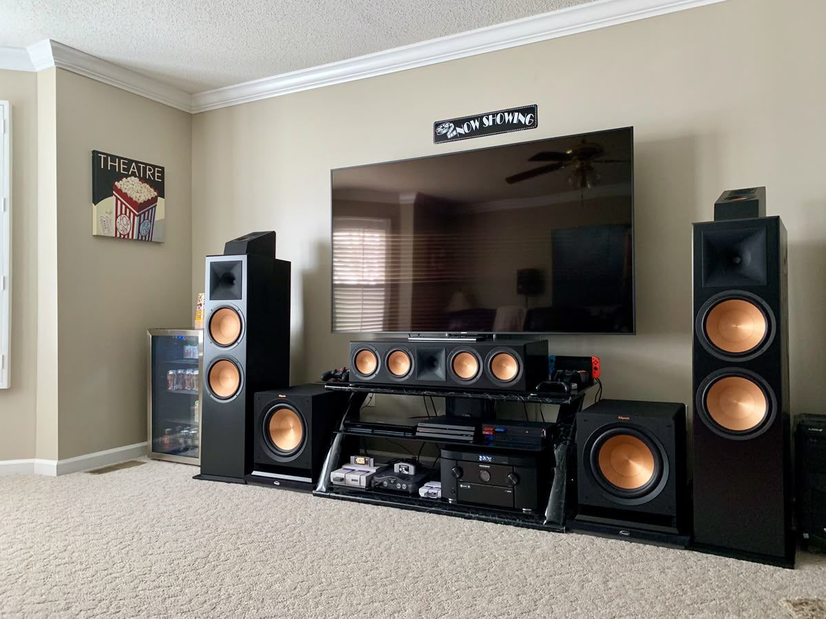Large flat screen TV wall mounted above a media console in a home theater room, flanked by Klipsch tower speakers with copper cones, a center channel and two subwoofers
